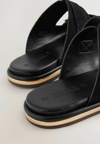 Sandales noires à glissière avec une finition lisse, accent doré sur la semelle et des sangles en tissu doux. Présente un logo de la marque sur le pied.