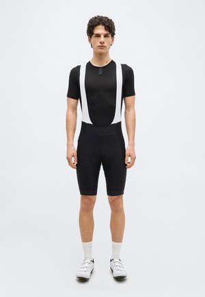 Jeune homme portant un cuissard de cyclisme noir avec des bretelles blanches, un maillot noir à manches courtes, des chaussettes blanches et des chaussures de cyclisme blanches, debout.