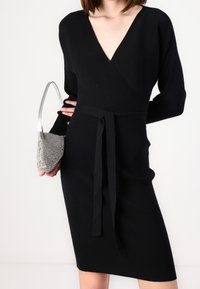 Svart ribbad stickad klänning med djup V-ringning, långa ärmar och ett knytbälte. Åtföljd av en silverfärgad metallhandväska med ett kort handtag.