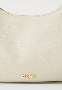 Beige bőr kézitáska pehely textúrával, ívelt formával és arany "DKNY" logóval az elején. Öltögetett részletek a széleken.
