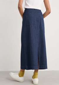 Džínsová maxi sukně tmavě modré barvy se dvěma zadními kapsami a bočním rozparkem. Nosit s bílými teniskami a žlutými žebrovanými ponožkami.