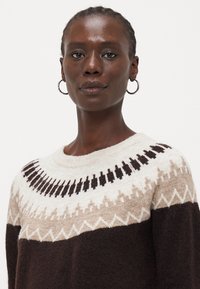 Strikkegenser med rund hals, som har et geometrisk mønster i krem, beige og mørkebrunt. Kjølig og myk tekstur med lange ermer.