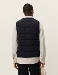 Μαύρο φουσκωτό γιλέκο με καπιτονέ σχέδιο, στρογγυλή λαιμόκοψη και χωρίς ορατές κλειδώματα. Φοριέται πάνω από ένα ανοιχτόχρωμο πουκάμισο.