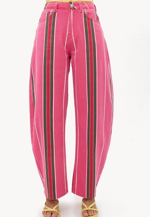Pantalon rose ample avec des rayures verticales rouges, vertes et blanches, porté avec des sandales jaunes à brides sur fond blanc.