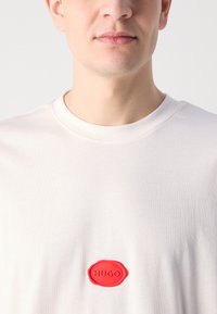 Hvid bomulds t-shirt med en rund rød gummipatch, der har "HUGO" præget i midten, glat tekstur og rund hals design.