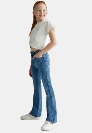 T-shirt bianco corto con dettaglio annodato sul davanti, abbinato a jeans a zampa d'elefante blu e sneakers bianche. Tessuto morbido e vestibilità casual.