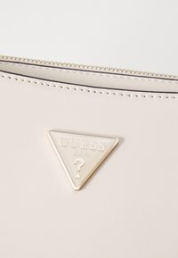Borsa a tracolla sintetica beige con logo metallico triangolare con la scritta "GUESS" e "U.S.A." in rilievo. Chiusura con zip e dettagli di cucitura a contrasto.