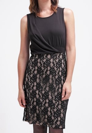 Femme portant un haut noir sans manches et une jupe beige longueur genou avec une superposition en dentelle florale noire, debout devant un fond blanc.