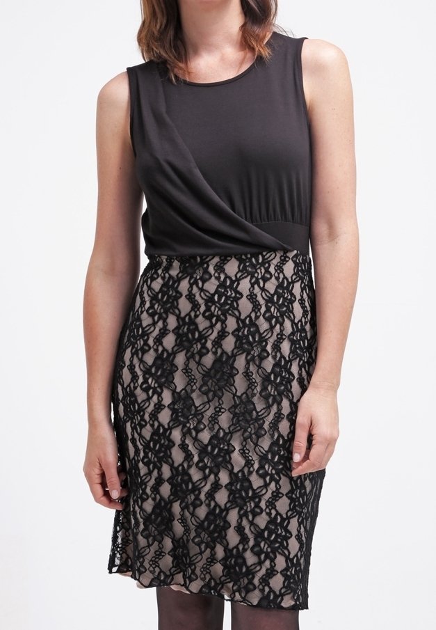 Femme portant un haut noir sans manches et une jupe beige longueur genou avec une superposition en dentelle florale noire, debout devant un fond blanc.