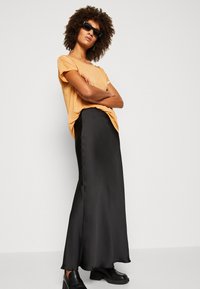 Svart satin maxi skjørt kombinert med en metallisk oransje t-skjorte med korte ermer. Modellen har på seg svarte solbriller og kraftige svarte sko.