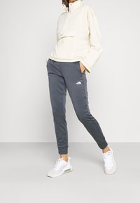 Osoba oblečená v krémovém pulovru Nike, šedých teplácích The North Face a bílých teniskách Nike, stojící s rukama v kapsách na bílém pozadí.