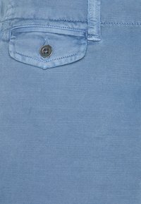 Tissu en denim bleu clair en gros plan montrant une petite poche avec un rabat sécurisé par un seul bouton foncé et des coutures diagonales visibles.