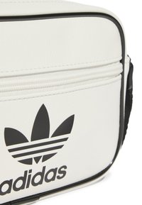 Borsa sintetica bianca con accenti neri, caratterizzata da un prominente logo Adidas nero, chiusura con zip e superficie testurizzata.