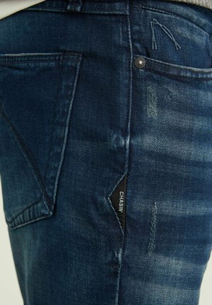 Tmavě modré džíny z denimu s výrazným zesvětlením, stříbrným knoflíkem a černou nálepkou na boční kapse.