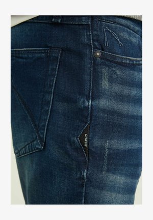 Tumedat sinise denimi teksad, millel on iseloomulik fadi muster, hõbedane nööpide detail ja must silt küljetaskul.