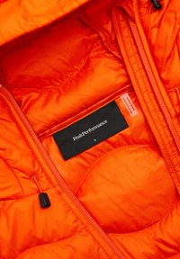 Helle orange isolierte Jacke mit Innenfutter aus Rautenstepp, ausgestattet mit einem schwarzen Etikett mit "PeakPerformance" und der Größe "S", sowie einem sichtbaren Reißverschluss.