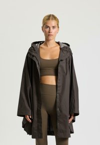 Cappotto impermeabile marrone, lungo fino al ginocchio, con cappuccio, bottoni a pressione e fodera grigia, indossato sopra un reggiseno sportivo marrone e leggins.