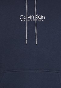 Felpa blu navy con cappuccio e lacci, caratterizzata da un design a pattern. La parte anteriore presenta un testo bianco che riporta "Calvin Klein" e coordinate geografiche.