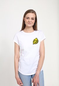 Bílý bavlněný tričko s ohrnutými rukávy, se žlutým a černým logem Avengers na levé hrudi. Nosené se světle modrými džínami.