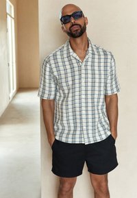Camicia a maniche corte fantasia in bianco e blu navy con vestibilità rilassata, abbinata a pantaloncini neri. Tessuto liscio con motivo a quadretti.