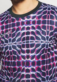 Krátký rukáv košile ze syntetického materiálu s živým abstraktním vzorem v růžové, bílé a modré barvě na tmavém pozadí, s černým kulatým výstřihem.