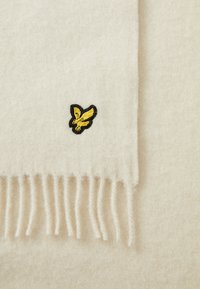 Écharpe en laine de couleur crème avec des extrémités à franges et un logo d'oiseau brodé noir et jaune. Texture douce avec une finition lisse.