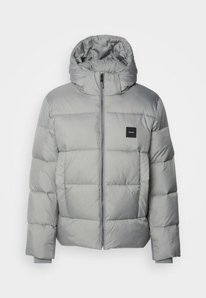Doudoune grise avec capuche, dotée d'une fermeture éclair, au design matelassé et d'un petit patch logo noir sur la poitrine gauche. Texture lisse.