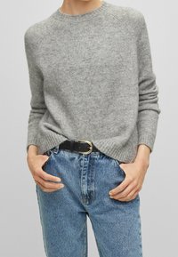 Ljust grått ulltröja med ribbade ärmslut och nederkant, buren över högmidjade blå denimjeans med ett svart bälte som har ett guldspänne.