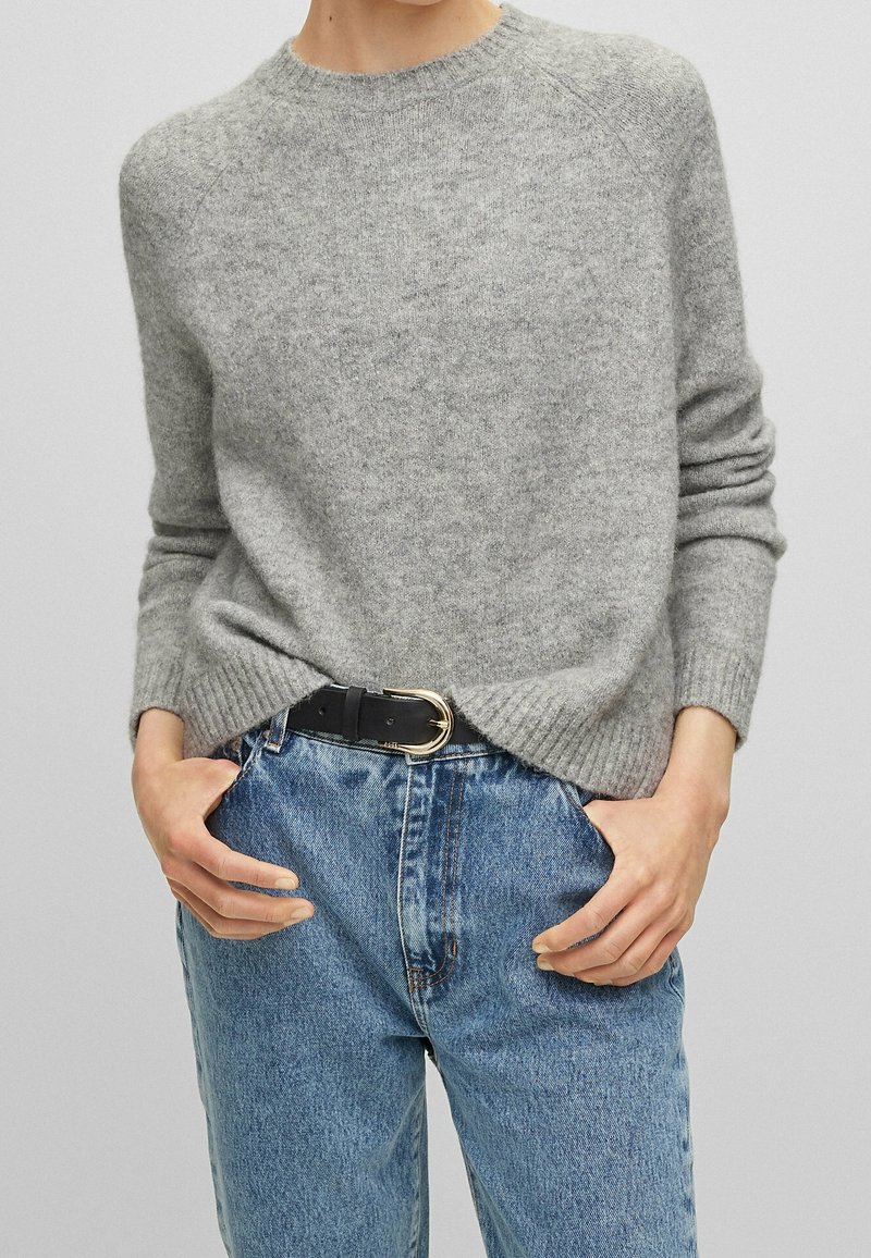 Ljust grått ulltröja med ribbade ärmslut och nederkant, buren över högmidjade blå denimjeans med ett svart bälte som har ett guldspänne.