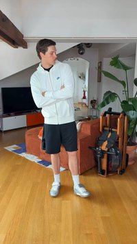 Jeune homme en sweat à capuche blanc et short noir, debout dans le salon, les bras croisés près d'un canapé orange et d'un rameur en bois à côté d'une plante.