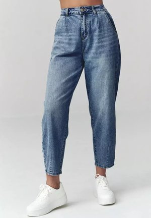 Niebieskie dżinsy o wysokim stanie i zwężanych nogawkach, noszone z białymi sneakersami na platformie, skrócone przy kostkach na jednolitym jasnym tle.