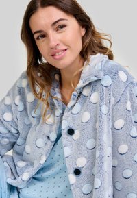 Abrigo de piel sintética azul claro con lunares blancos y azul claro, mangas largas y botones negros. Llevado sobre una blusa azul claro con lunares.