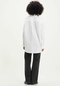 Chemise en coton blanc avec un ourlet arrondi et des poignets à un bouton, assortie à un pantalon large gris foncé. Le dos présente un détail de couture en yoke.