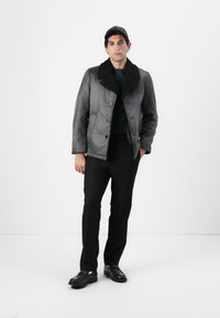 Giacca di shearling grigia con colletto nero texturizzato, due tasche frontali e bottoni. Abbinata a pantaloni neri e scarpe lucide.