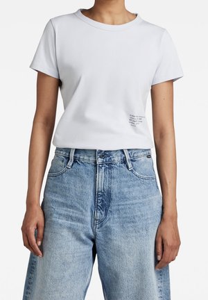 Osoba ubrana w prosty biały t-shirt z krótkim rękawem, wsunięty w jasnoniebieskie jeansy z wysokim stanem, stojąca na białym tle.