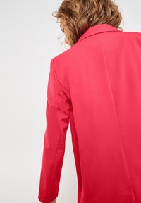 Červený blazer s přizpůsobeným střihem, dlouhými rukávy, strukturovaným límcem a hladkou texturou látky. Pohled z zad zdůrazňuje čisté linie.