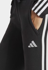 Zwarte sportbroek met witte strepen, heeft een gladde structuur, elastische tailleband en een klein zilver logo aan de rechterkant.