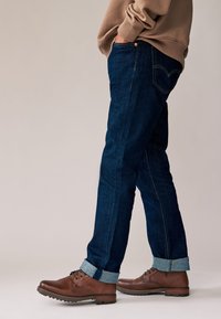 Tmavomodré džínsy z denimu s rovnými nohavicami, ohrnutými lemami a žltým prešívaním. Nosia sa s hnedými koženými topánkami a béžovou mikinou.