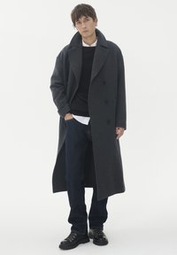 Lång mörkgrå kappa i ullblandning med bred krage, dubbelknäppt front och sidofickor, i kombination med svart tröja, vit skjorta och mörka jeans.