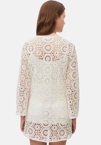 Weißes gehäkeltes Kleid mit langen Ärmeln, das hexagonale und florale Muster aufweist und ein komplexes Spitzenmuster sowie einen strukturierten Stoff zeigt.