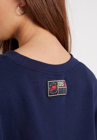 Mörkblå sweatshirt med en texturerad yta, som har en svart etikett med röda och gröna accenter, placerad på baksidan nära halsringningen.