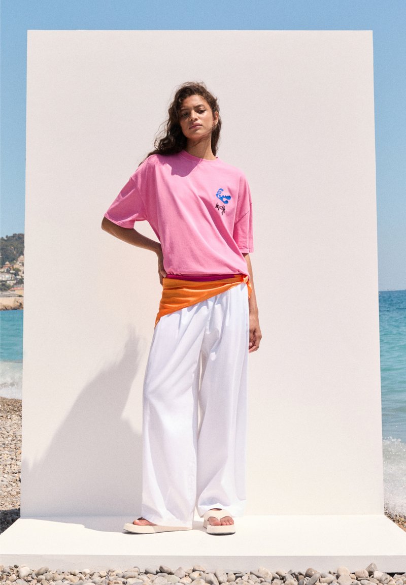 Lyserød oversize t-shirt med blå og sort grafisk design, parret med hvide brede bukser og en orange sjaal. Sandaler fuldender outfittet.