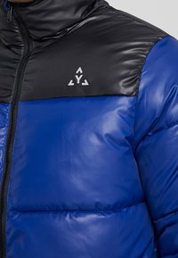 Blå puffjacka med en svart, glänsande övre del, en framdragkedja och en logotyp p å bröstet; slät textur med ett kviltat mönster.