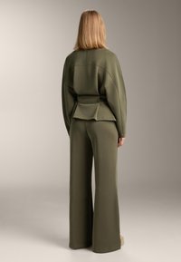 Olívazöld összeillő szett, bő szabású felsővel, deréknál övvel és bőszárú nadrággal, puha, texturált anyagból készült.