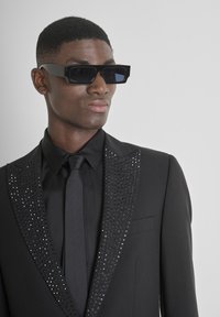 Zwart pak met glinsterende paillettenaccenten op de revers, gecombineerd met een bijpassende stropdas en hoekige zwarte zonnebril. Soepele textuur, nauwsluitend ontwerp.