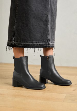 Schwarze Leder-Ankle-Boots mit abgerundeter Spitze und Blockabsatz. Mit einem seitlichen Reißverschluss und glatter Textur. Getragen mit einem ausgefransten Denim-Rock.