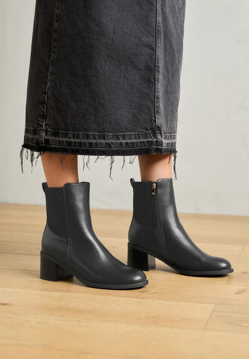 Stivaletti in pelle nera con punta arrotondata e tacco a blocco. Presentano una zip laterale e una texture liscia. Indossati con una gonna di jeans sfilacciata.