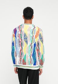 Άνδρας που φοράει πολυχρωμο πλεκτό πουλόβερ με σχέδια και κείμενο, στέκεται με την πλάτη προς την κάμερα σε λευκό φόντο.