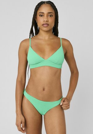 Femme aux longues tresses portant un bikini vert clair, debout devant un fond gris uni, tenant le côté du bas de bikini.