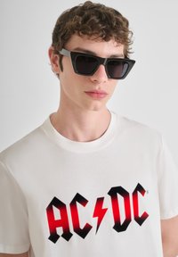 Camiseta de algodón blanca con un logo de AC/DC en negro y rojo, acompañada de unas gafas de sol negras y angulares con detalles en gris.